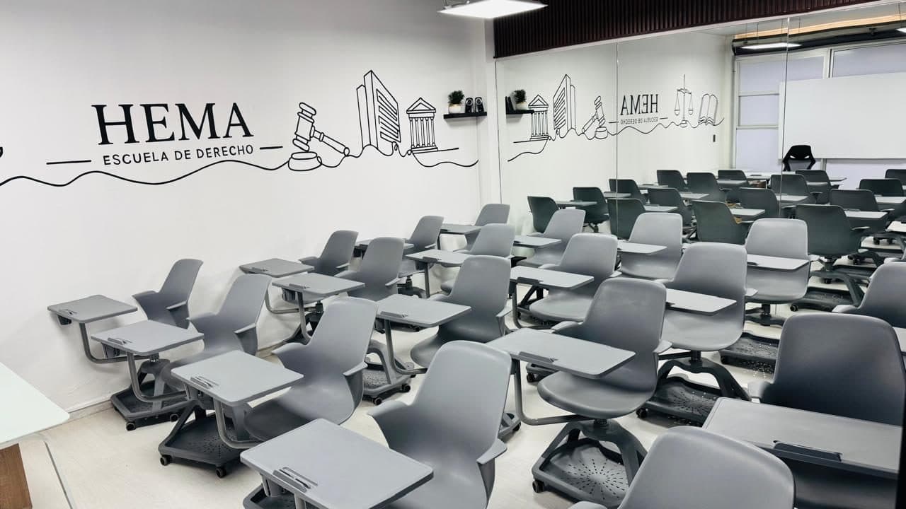 Aula del Instituto HEMA con filas de butacas con paleta sobre ruedas junto a la pared del mural de línea «HEMA · Escuela de Derecho», donde están dibujados un mazo de juez y un edificio de columnas.