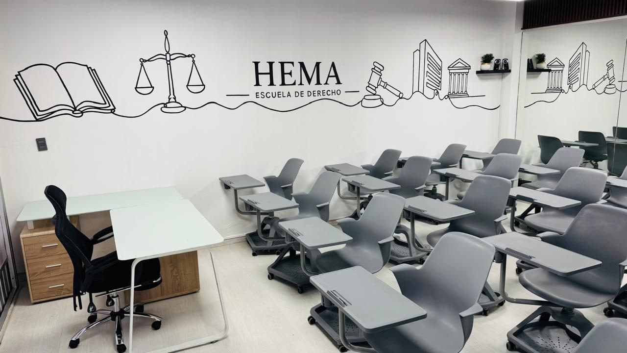 Frente de la misma aula: el escritorio y la silla del docente junto al mural con un libro abierto y una balanza de la justicia; a la derecha, las butacas de los estudiantes.